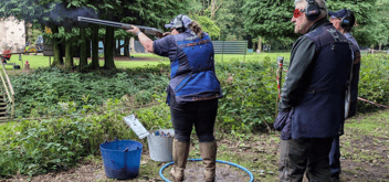 Emma Stacey. Woman firing shotgun in a shooting competition with two men in frame.